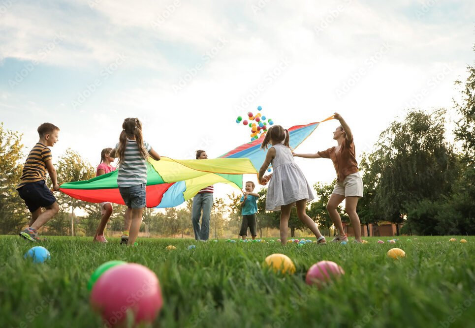 Group of children and teachers playing with rainbow playground parachute on green grass, low angle view. Summer camp activity
